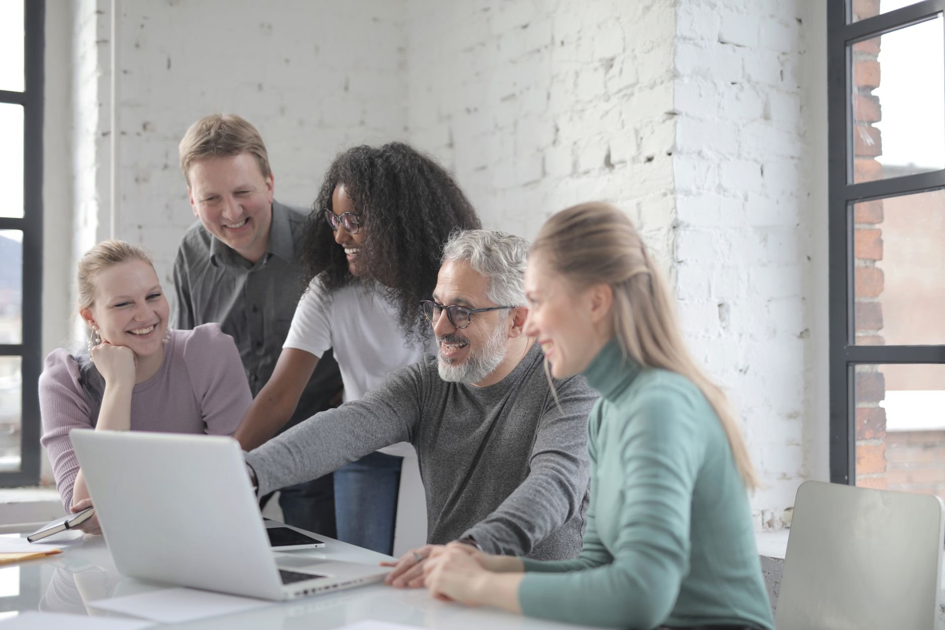 Positive diverse coworkers smiling and discussing details of project while working with laptop together in modern workspace